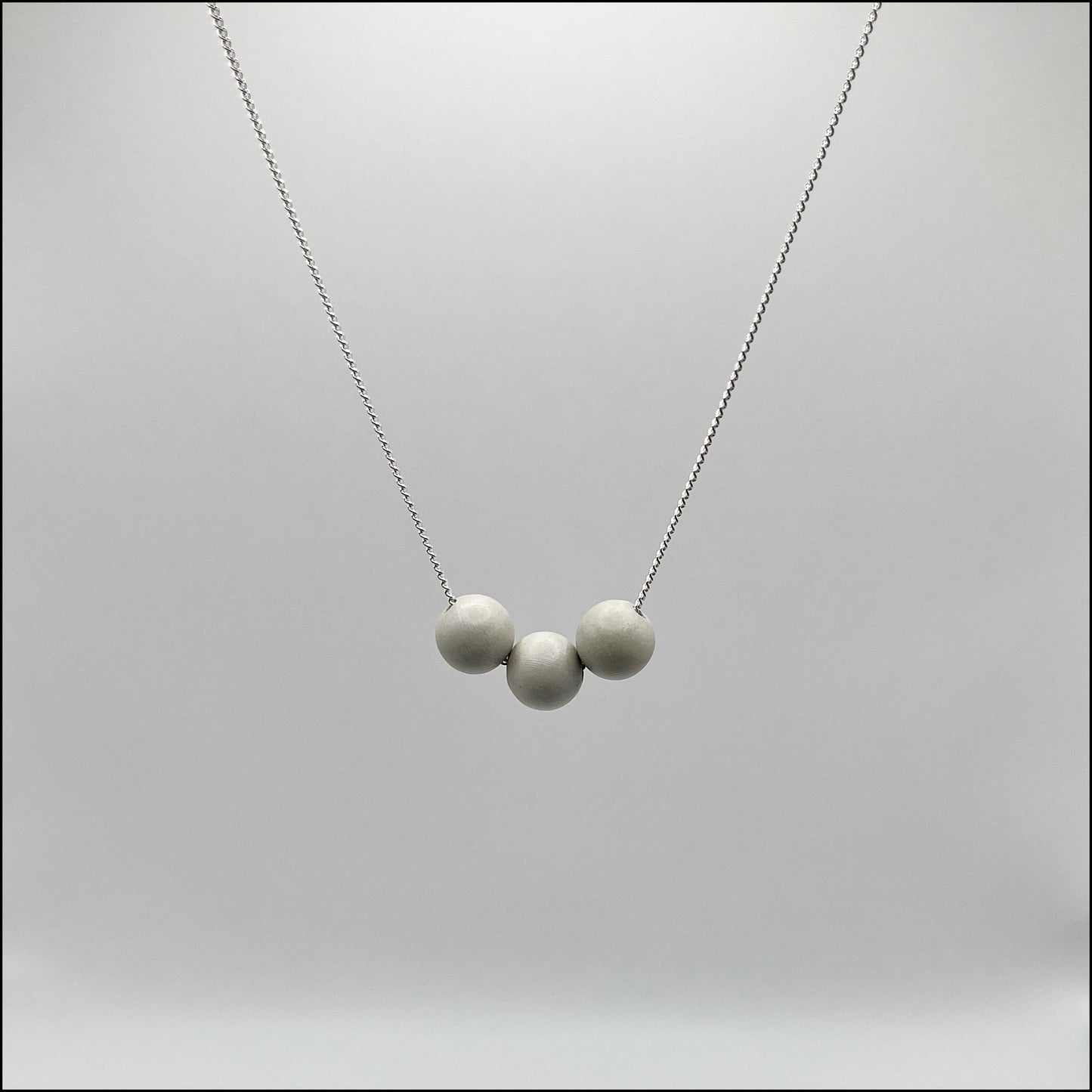 A silver curb chain necklace with three concrete spheres attached to it, displayed against a grey background.