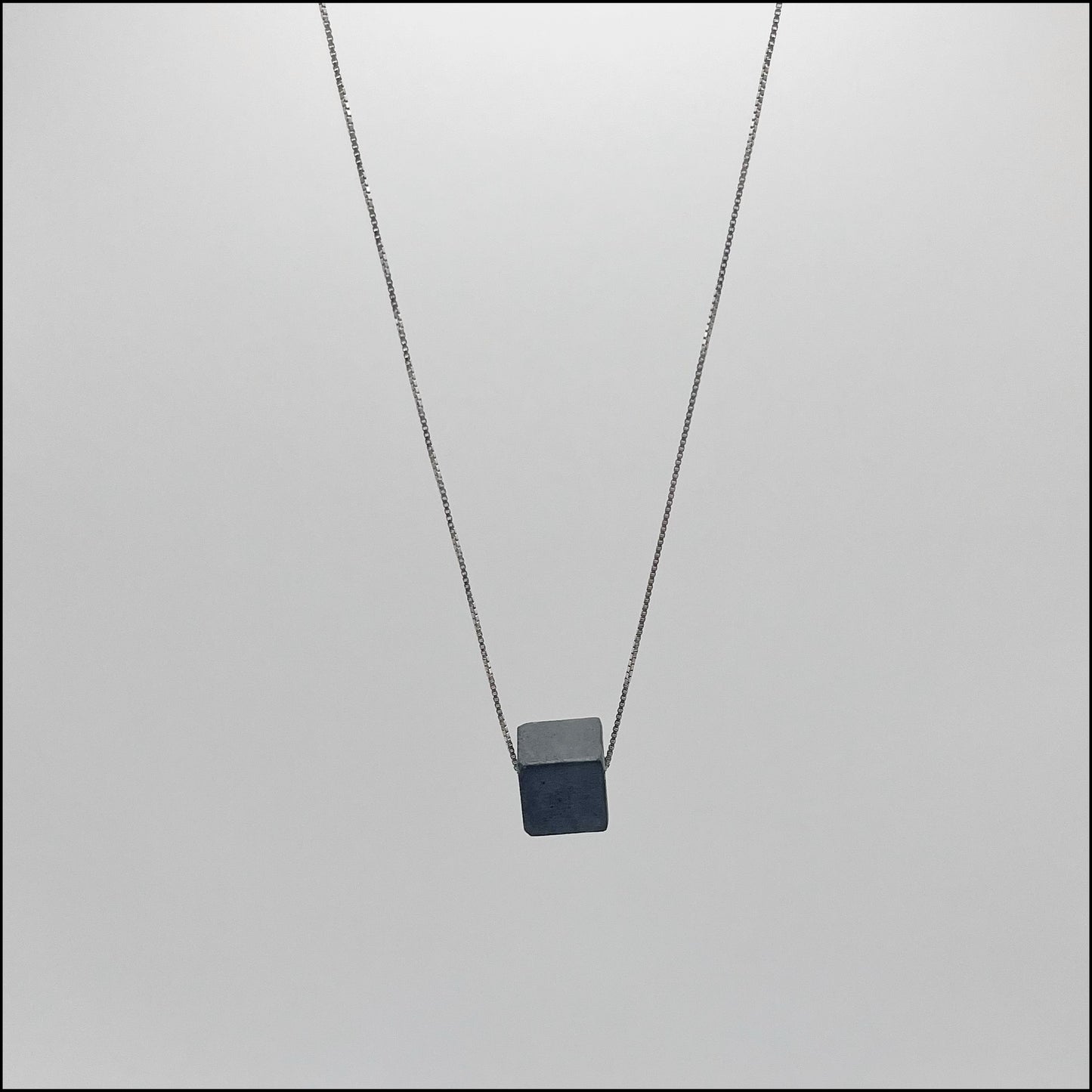 A silver box chain necklace with a teinted anthracite concrete cube pendant, displayed against a grey background