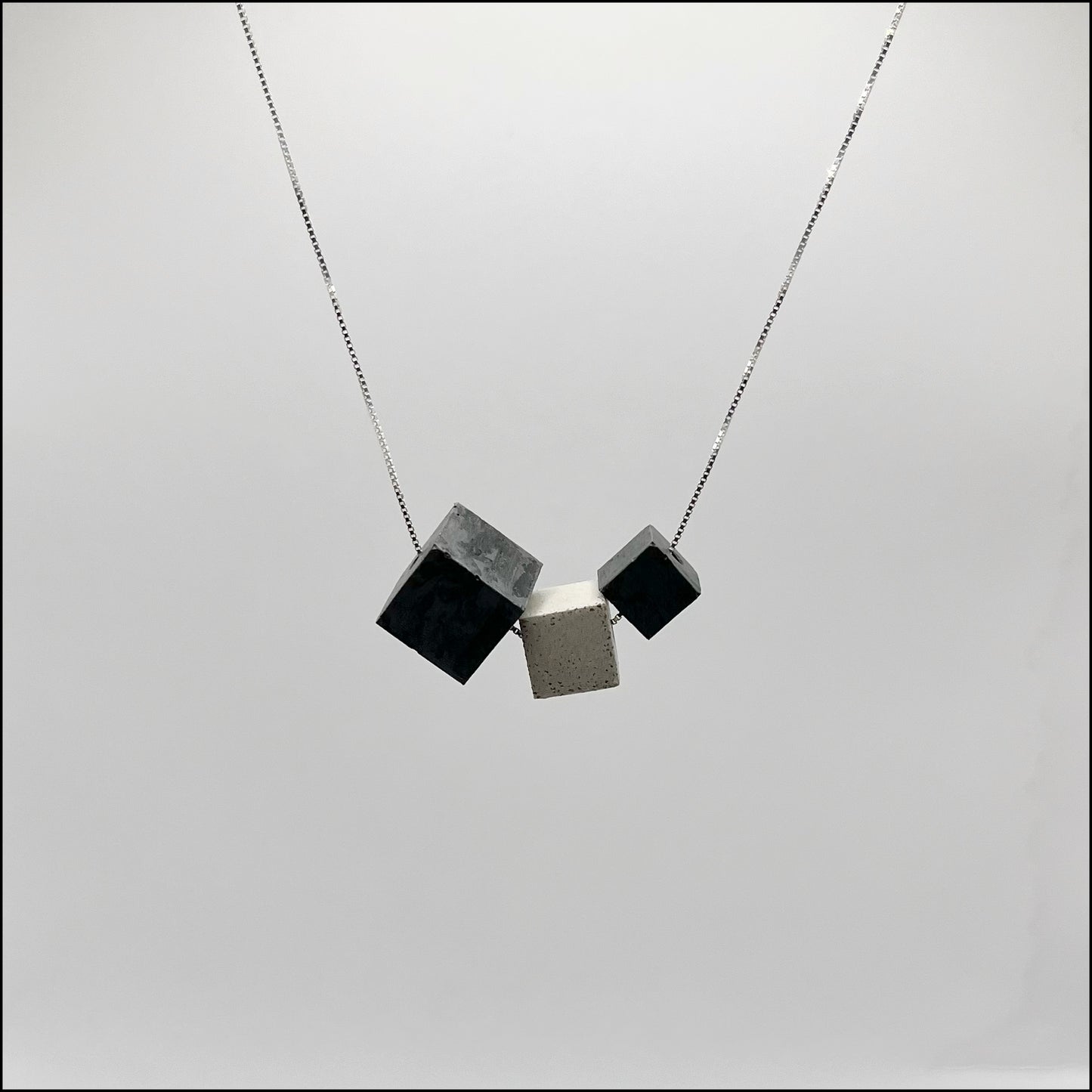 A silver chain necklace with three concrete cube pendants in varying sizes and colors, displayed against a grey background.