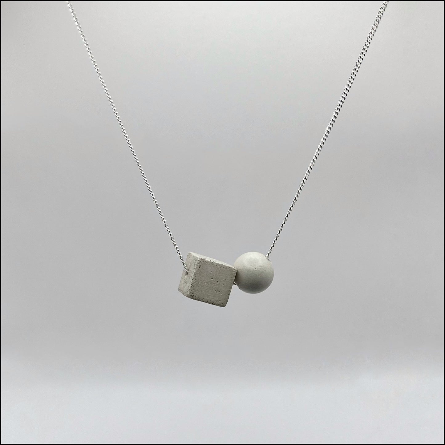 A silver curb chain necklace with a large concrete sphere and cube pendant attached to it, displayed against a grey background.