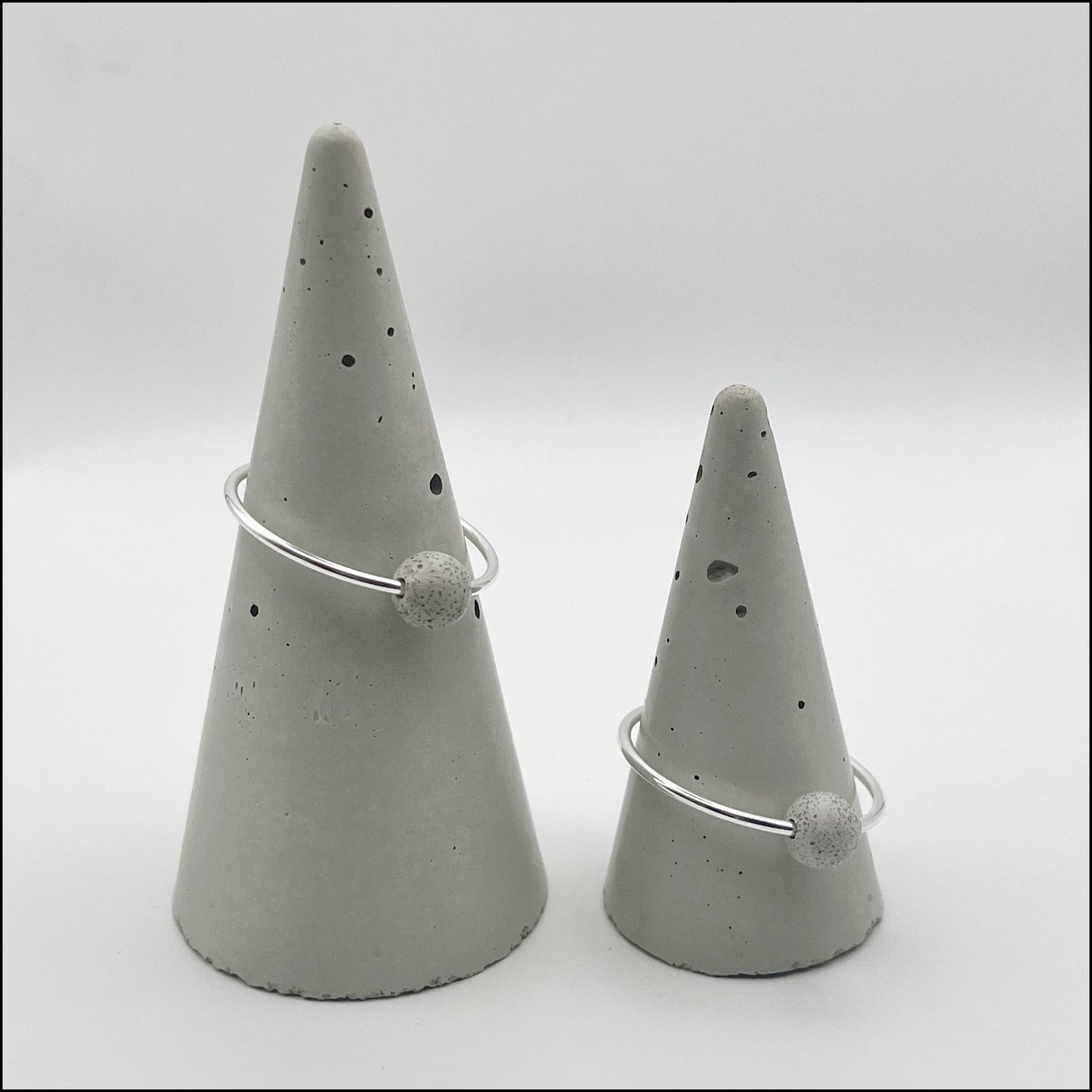 Two silver hoop earrings with handmade concrete spheres displayed on gray cone-shaped stands.